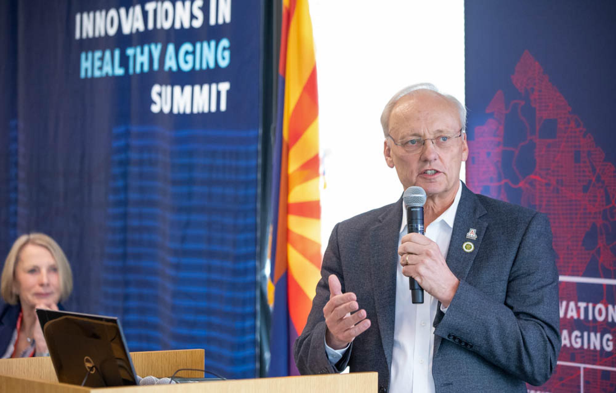 An older man wearing glasses and a suit speaks into a microphone at a podium in front of banners that say &quot;Innovations in Healthy Aging Summit.&quot;