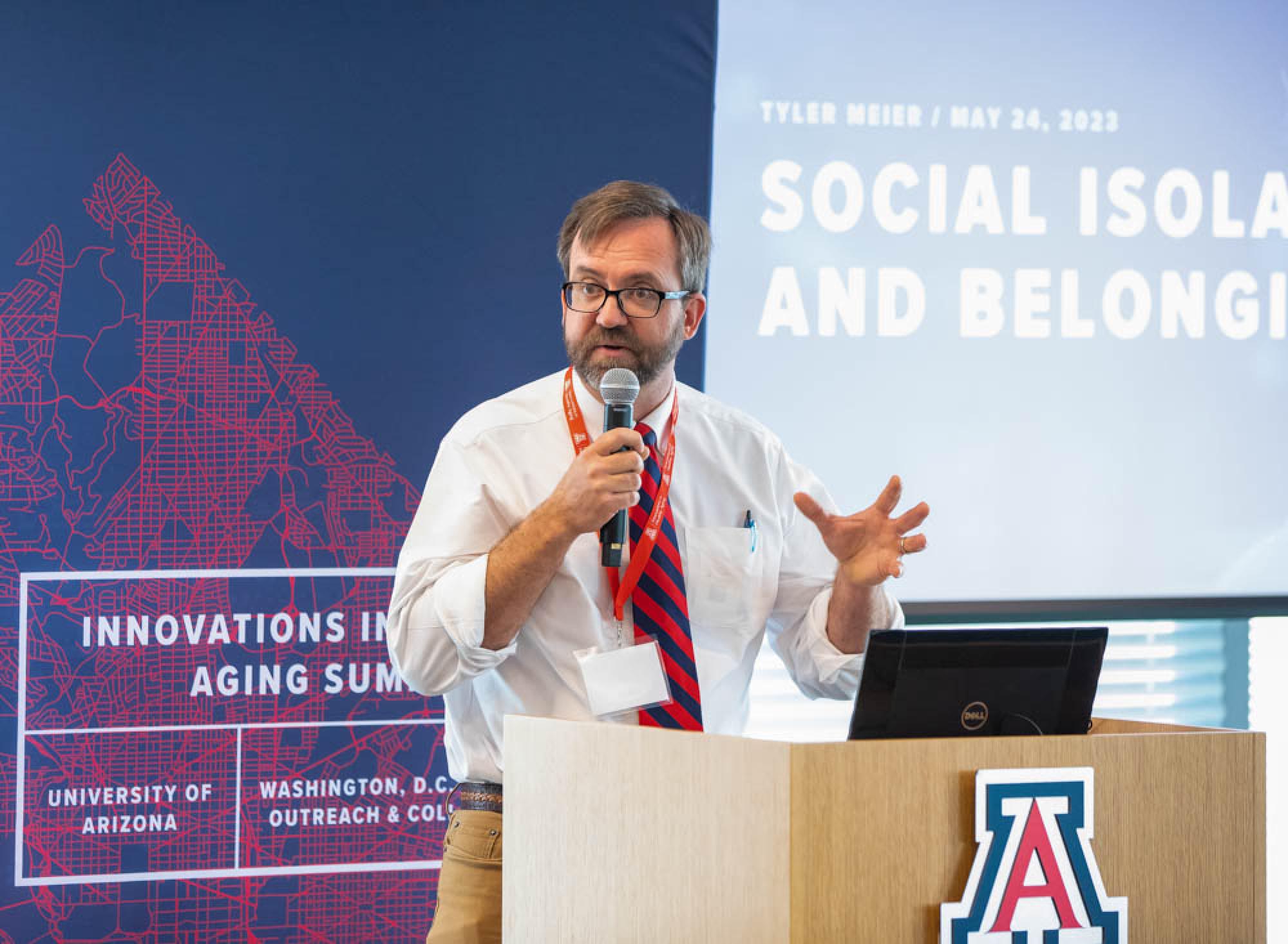 A brown-haired man speaks fervently into a microphone at a podium with the University of Arizona logo. A graphic displayed on a screen behind him reads &quot;Social Isolation and Belonging.&quot;