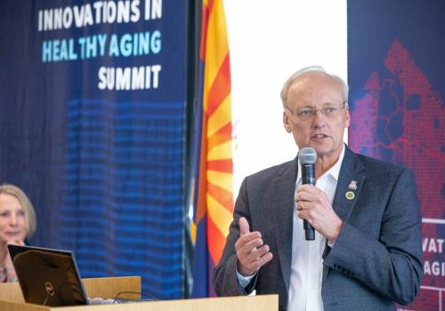 An older man wearing glasses and a suit speaks into a microphone at a podium in front of banners that say &quot;Innovations in Healthy Aging Summit.&quot;