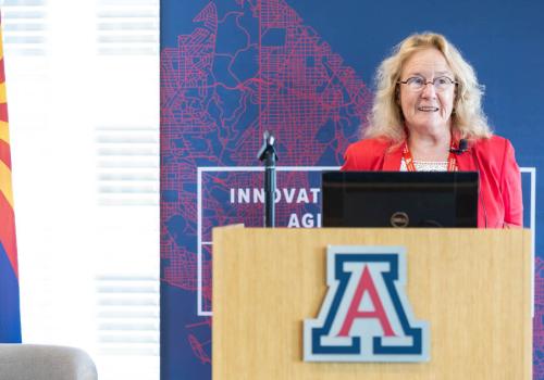 A woman wearing a bright red blazer stands at a podium with the University of Arizona logo.