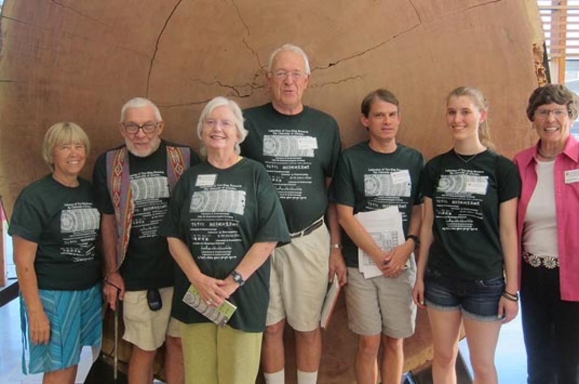 Group of men, women and students standing in front of a giant slice of tree displaying tree rings.